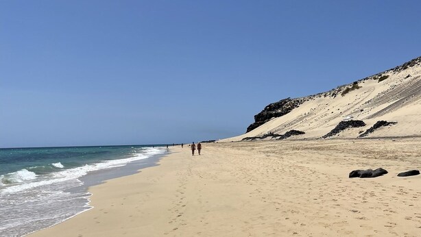 Une vaste plage de sable fin, parsemée d’empreintes menant à l’eau, une falaise rocheuse sur la droite et un ciel bleu azur.