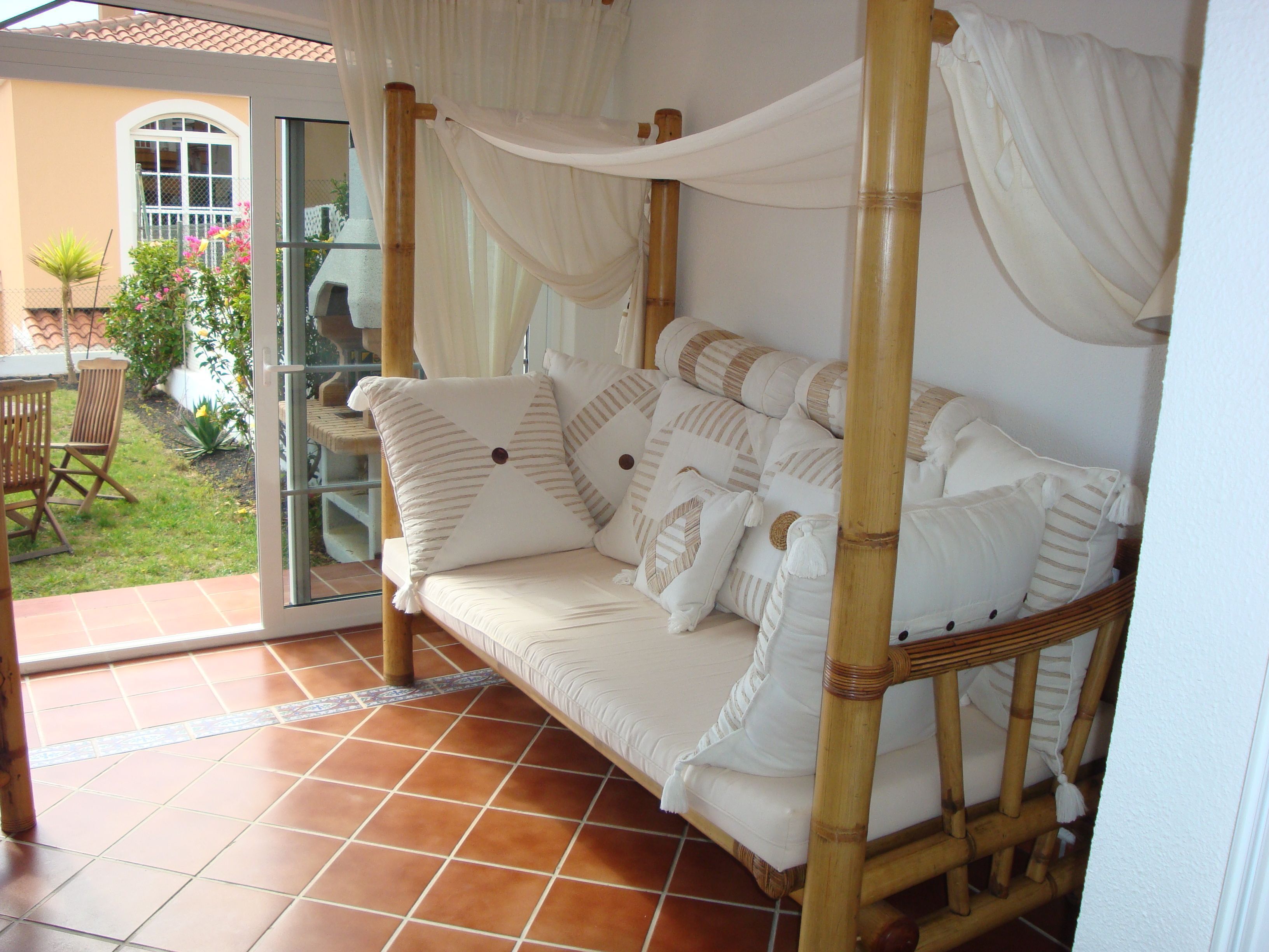 Una habitación con sofá de estructura de bambú, cojines blancos y una puerta de vidrio que da a un patio con una silla y plantas.