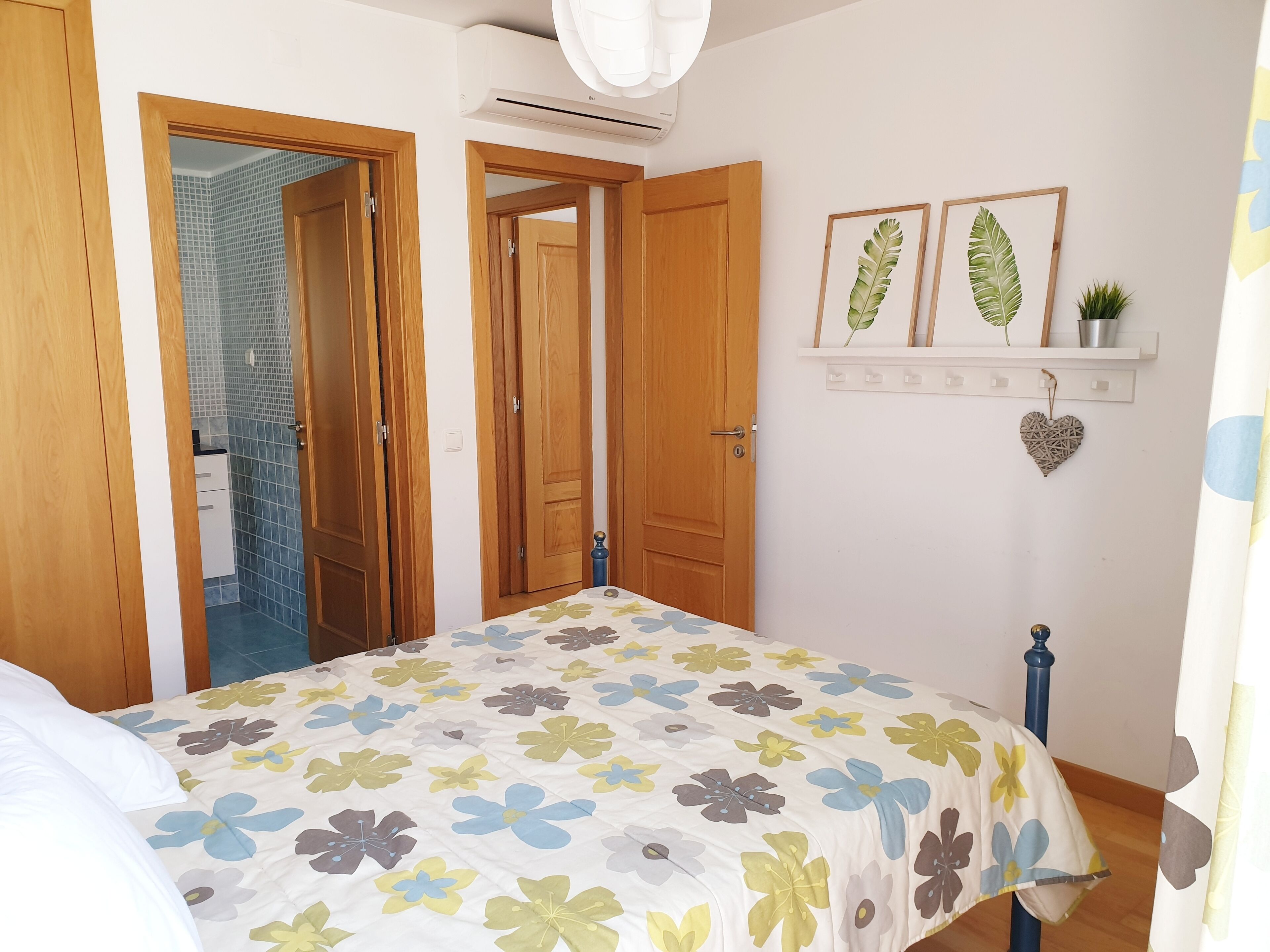 Un dormitorio con cubrecama floral, puertas de madera, un estante con adornos y un baño visible con azulejos azules.