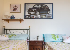 Une chambre avec deux lits, chacun recouvert d’une couvre-lit à motifs de cactus, une table de chevet avec une lampe et une étagère murale sur laquelle est posé un modèle de voilier.