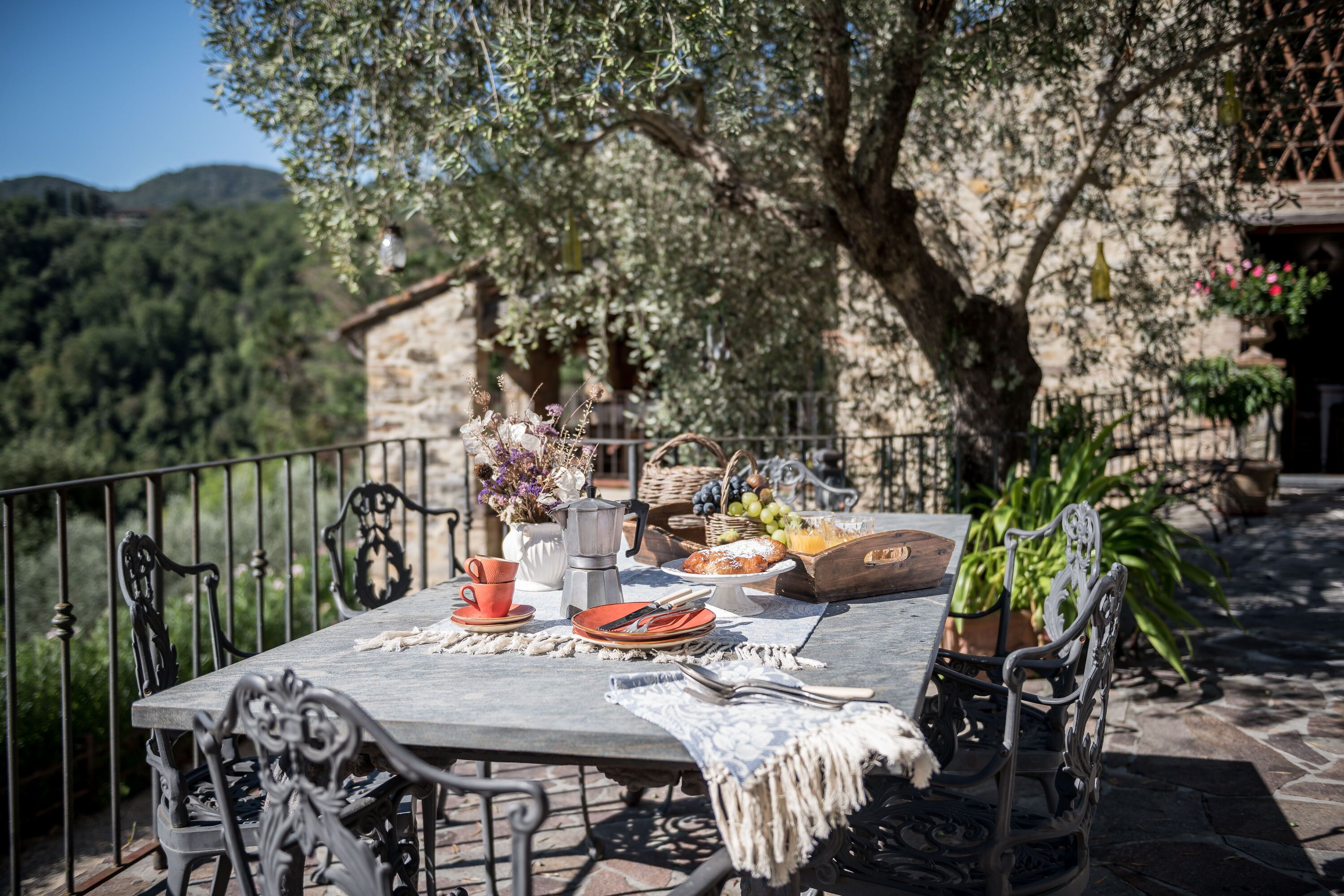 An outdoor dining table set with plates, cutlery, and a basket of bread, surrounded by ornate chairs and a rustic stone building in the background.