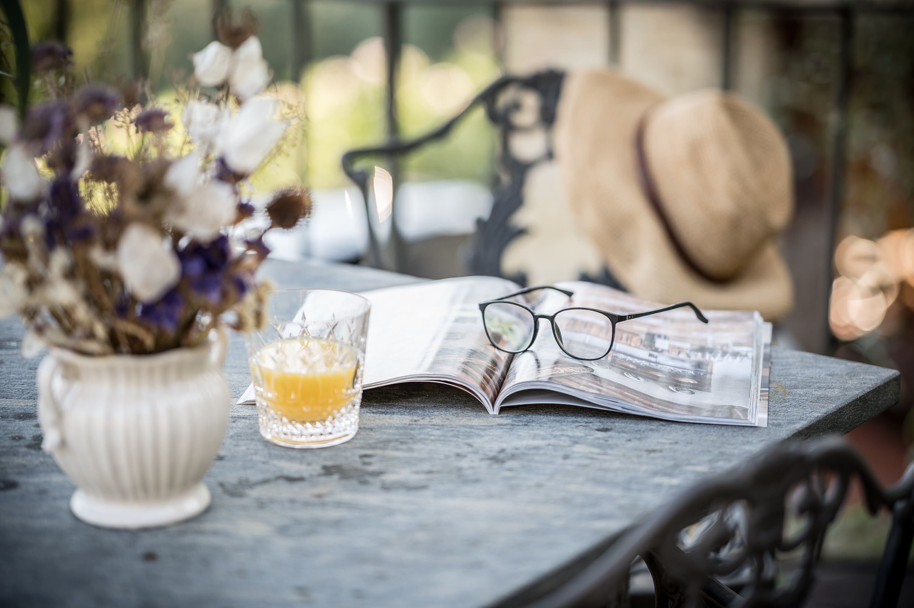 A table with a book, a pair of glasses, a glass of orange juice, and a vase of flowers.