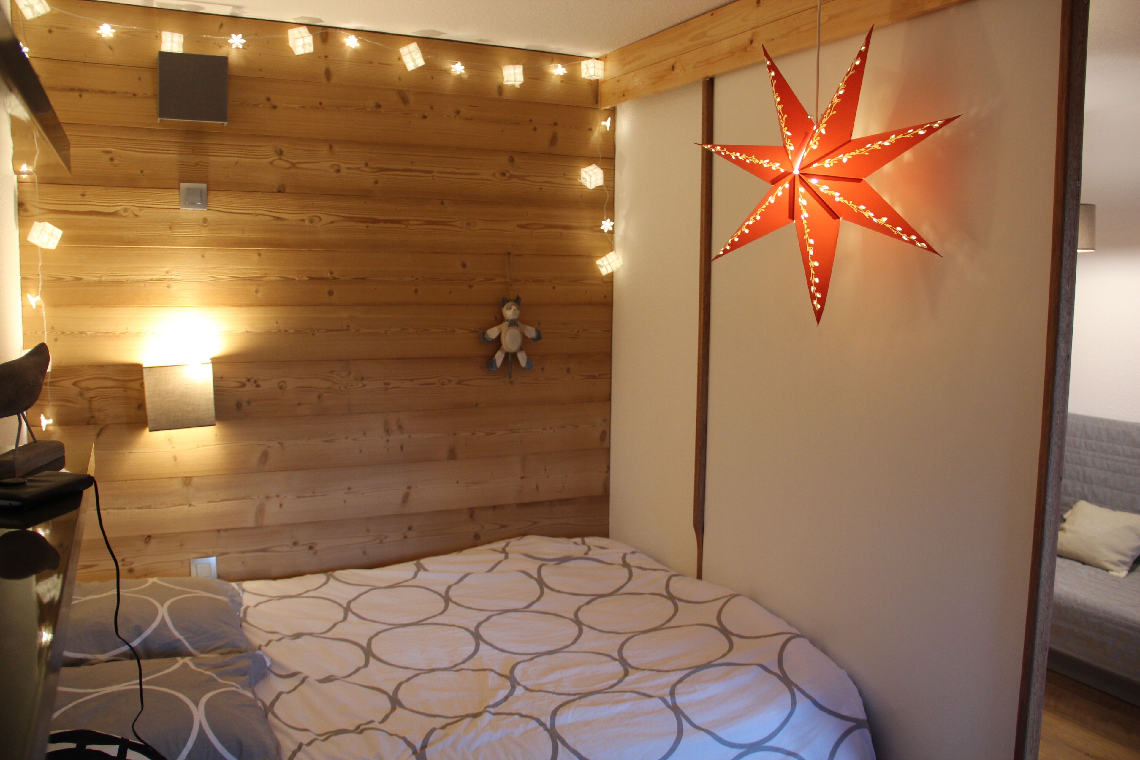 A cozy room with a wooden ceiling, a bed with a patterned bedspread, a wall-mounted star-shaped light, and a teddy bear decoration.