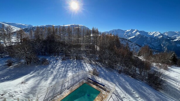 Ein Pool, umgeben von schneebedeckten Bäumen und Bergen unter blauem Himmel.