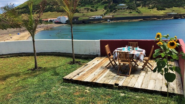 Une terrasse en bois avec une table et des chaises, donnant sur une plage et une étendue d’eau.
