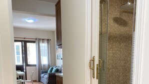 A hallway leading to a shower area with a glass door, wooden flooring, and a view into a living area with a sofa and a table.