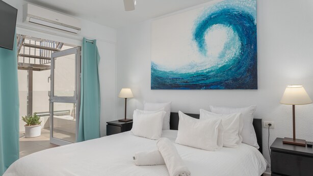 Une chambre d’hôtel avec un grand lit, du linge de lit blanc, un tableau représentant une vague bleue, une plante en pot et une porte coulissante donnant sur le balcon.