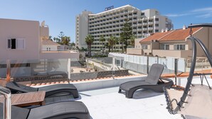 Une terrasse sur le toit, dotée de chaises longues et d’une balustrade en verre offrant une vue sur le paysage urbain.
