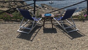 Eine Terrasse mit zwei blauen Liegestühlen, einem kleinen Tisch mit blauem Tablett und Blick auf das Meer und die felsige Küste.