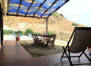 A covered patio with a table and chairs, a potted plant, and a view of a hilly landscape.