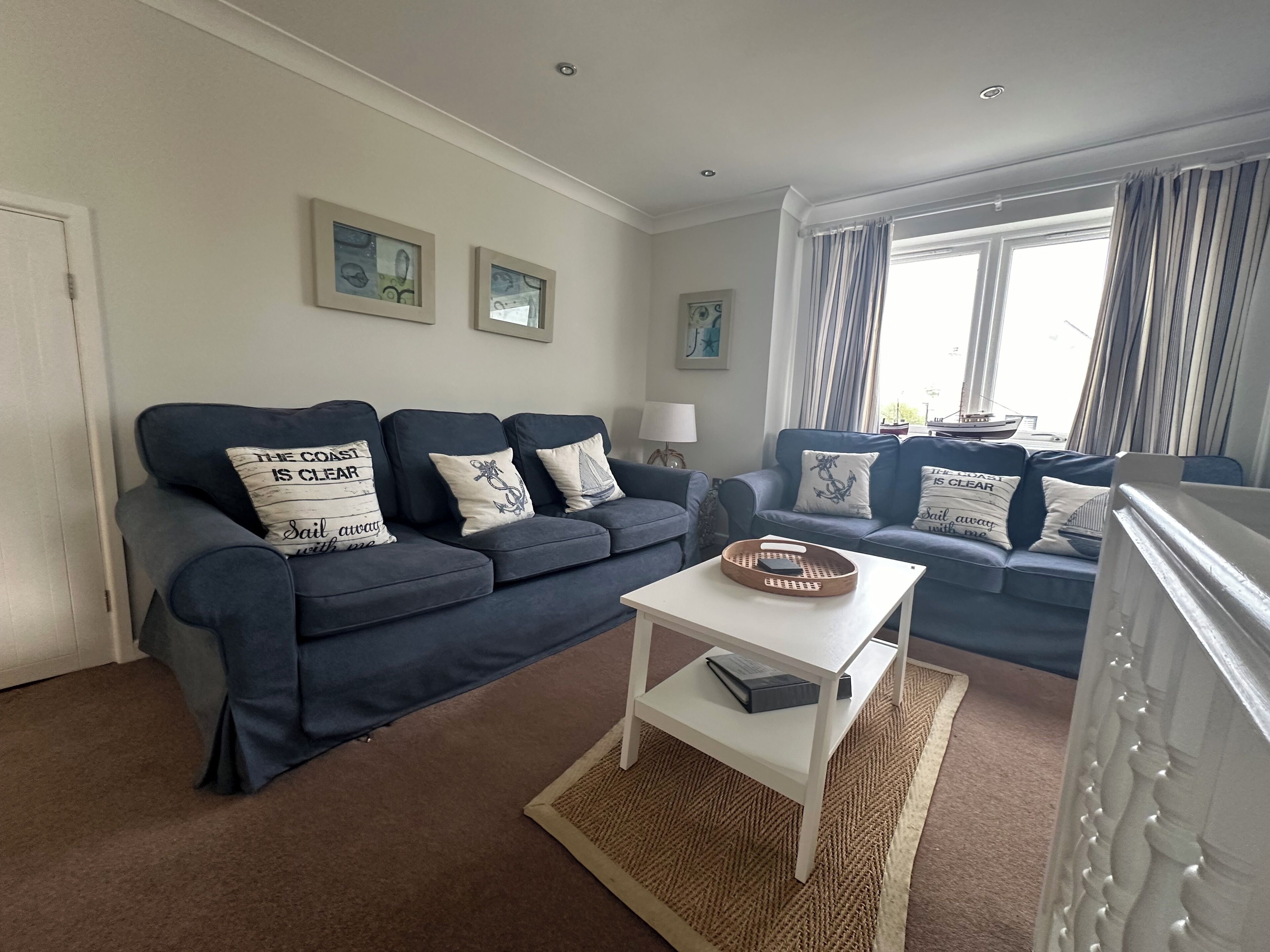 A living room with two dark blue sofas, a white coffee table, and a small white side table. There are framed pictures on the wall and a window with curtains.