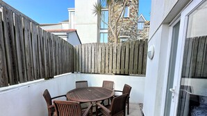 Une terrasse avec une table ronde et six chaises, entourée d’une clôture en bois et d’un mur blanc.