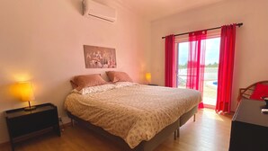 A bedroom with a bed, two bedside lamps, a painting on the wall, and a view through a window with red curtains. - Casa do Farol – Cozy Retreat with Special Rates in Low Season Algarve (Carvoeiro)