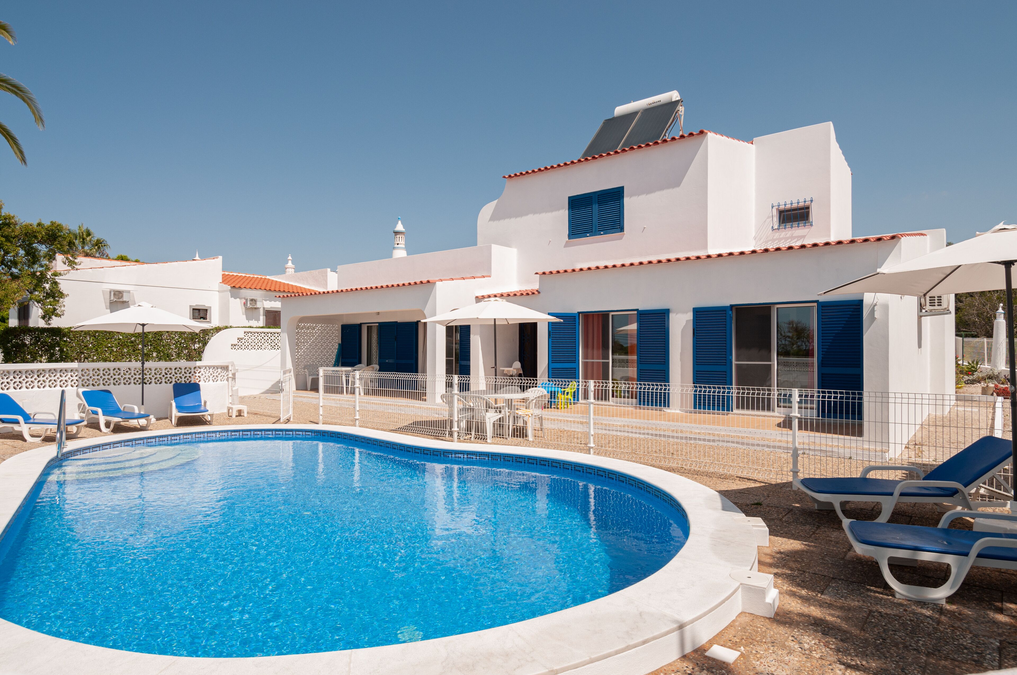 Una piscina con sillones de descanso, un edificio blanco con contraventanas azules y un cielo azul despejado.
