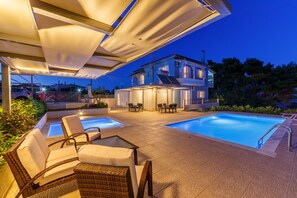 A poolside area with lounge chairs and a covered seating area, a large house in the background, and a clear blue sky. - Friends & Family House near the beach with two swimming pools (one for children) (Petalidi)