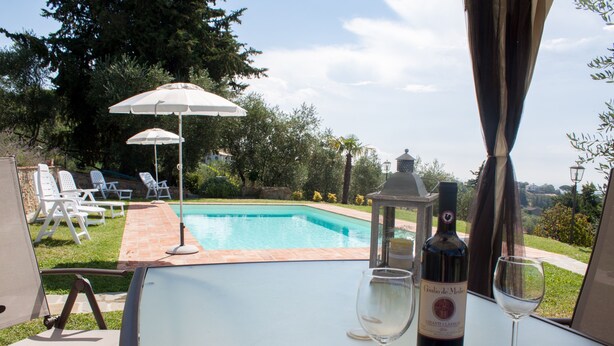 Ein Tisch am Pool mit einer Weinflasche und zwei Gläsern, mit Blick auf einen Swimmingpool mit Liegestühlen und Sonnenschirmen.