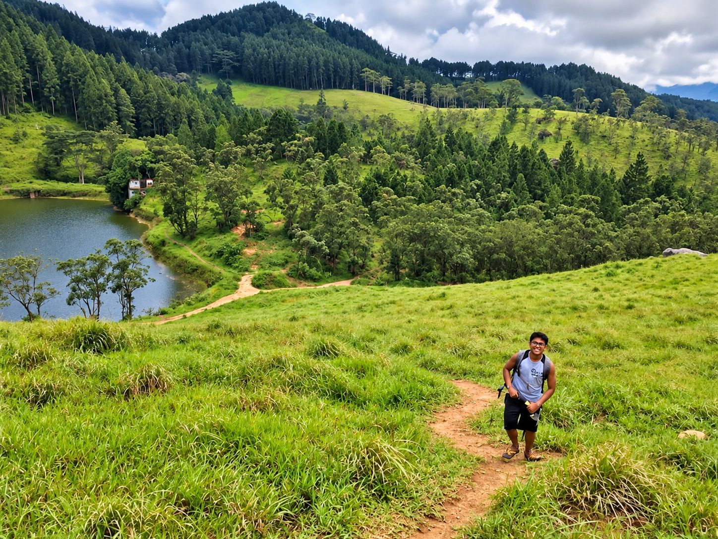A person standing on a dirt path in a grassy area with a lake and forested hills in the background.