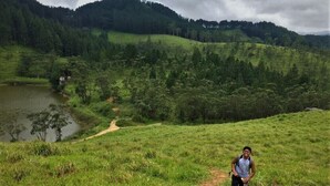 Une personne marchant sur un sentier de terre dans un champ d’herbe, avec un lac et des collines boisées en arrière-plan.