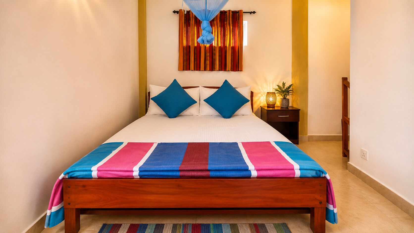 A bed with a colorful striped bedspread, a nightstand with a lamp, and a window with curtains.
