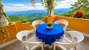 Ein runder Tisch mit blauer Tischdecke und weißen Korbstühlen auf einem Balkon mit Blick auf grüne Hügel.