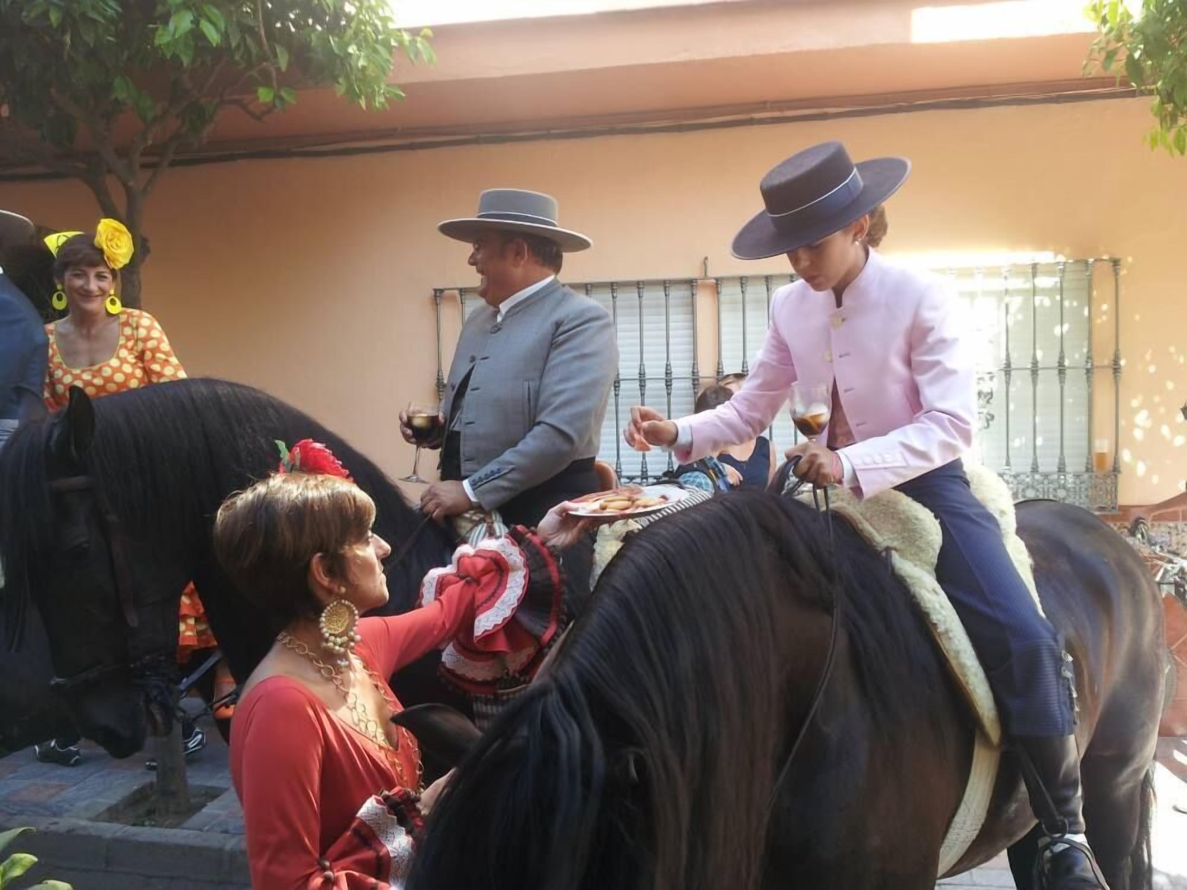 A group of people on horseback, some wearing hats, in an outdoor setting with a building in the background.