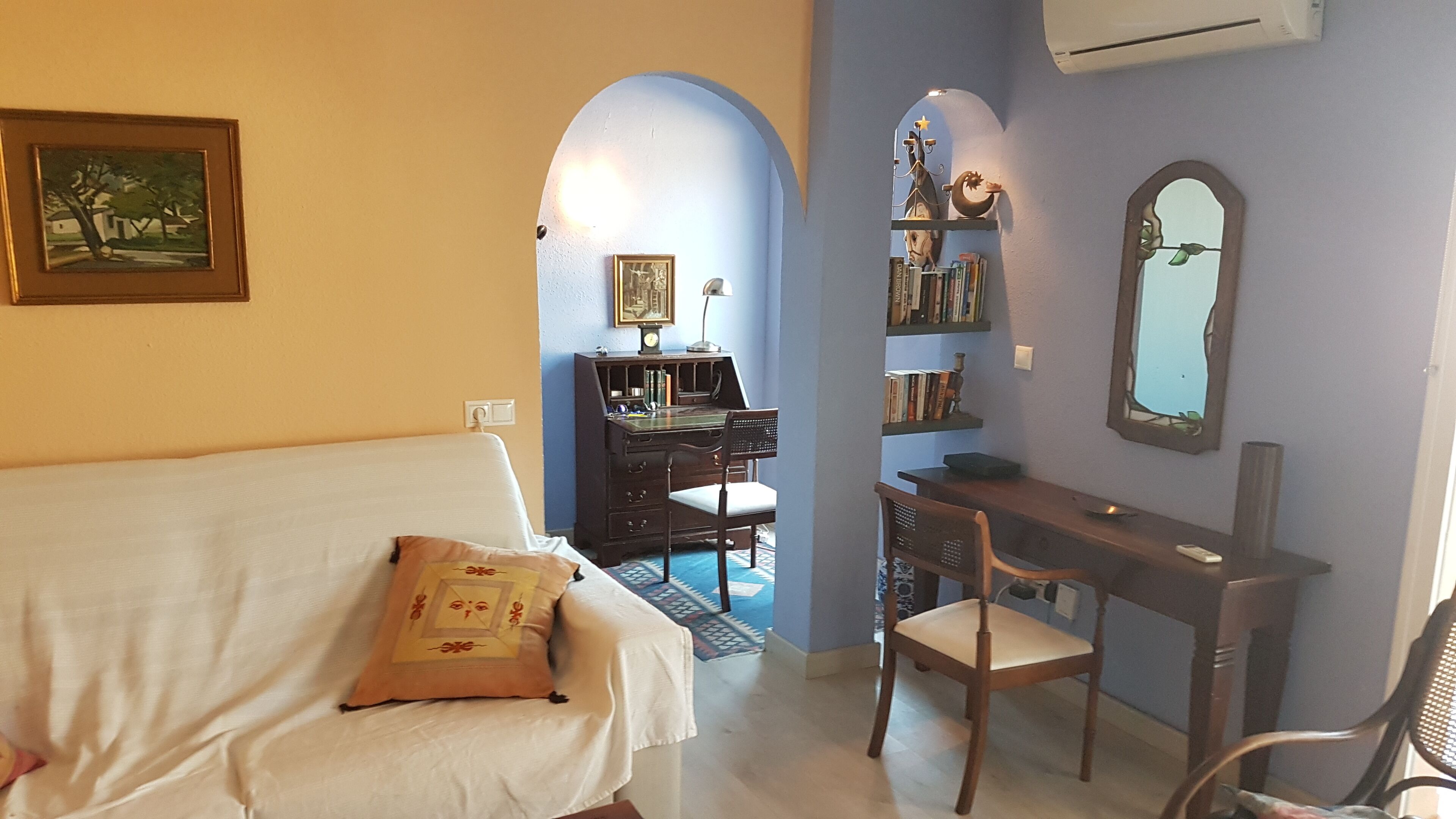 A room with a white sofa, a desk with a chair, a bookshelf, and a mirror on the wall.