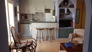 A compact kitchen with white cabinets, a tiled backsplash, and a central island. There are two wooden stools and a wicker chair. A small table with a blue folder is on the right. - Large sunny terrace, 6à m from the beach; large and comfortable (Fuengirola)