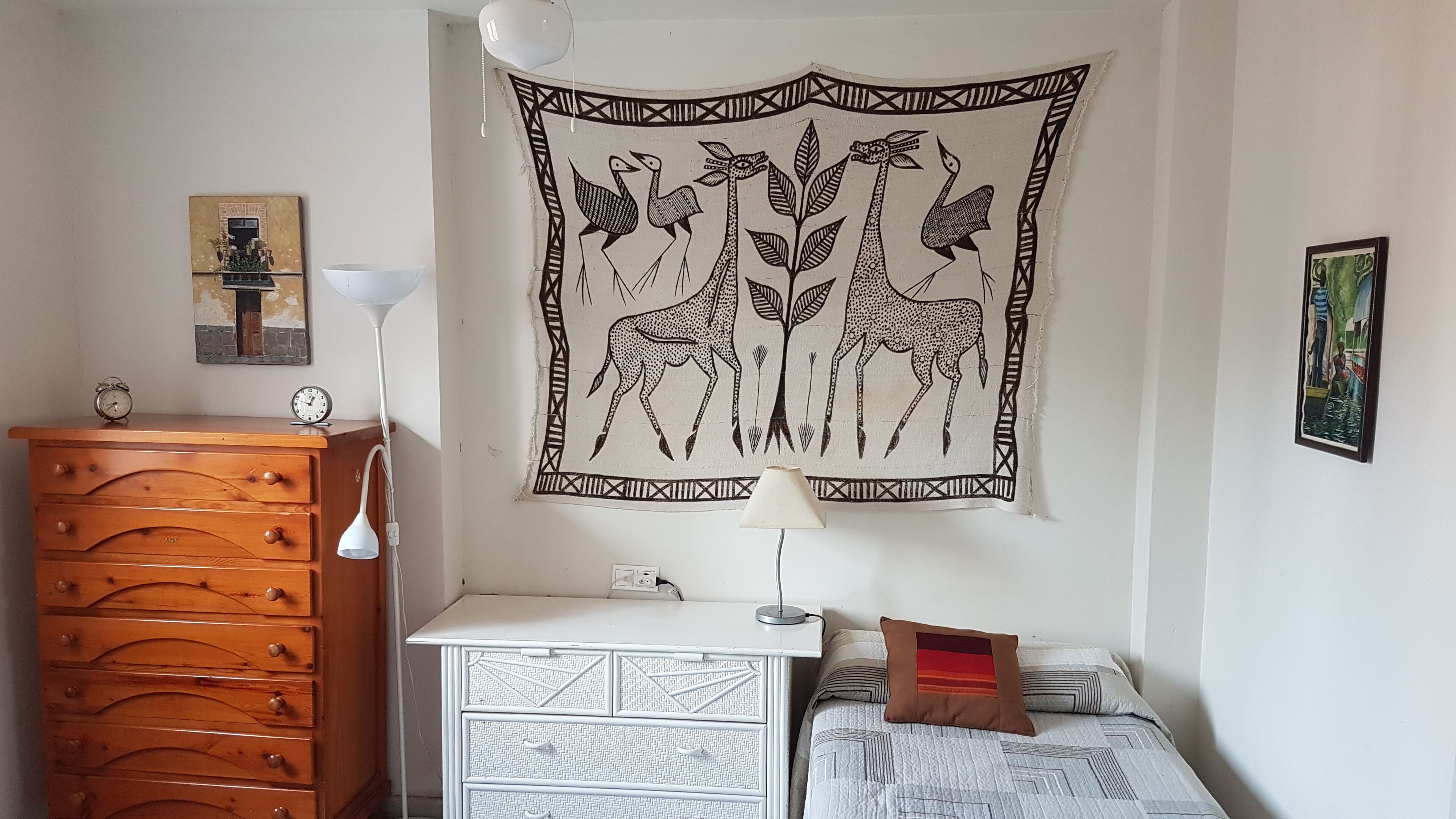 A bedroom with a wooden dresser, a white bedside table, a lamp, a bed with patterned bedding, and a tapestry with animal and plant designs on the wall.