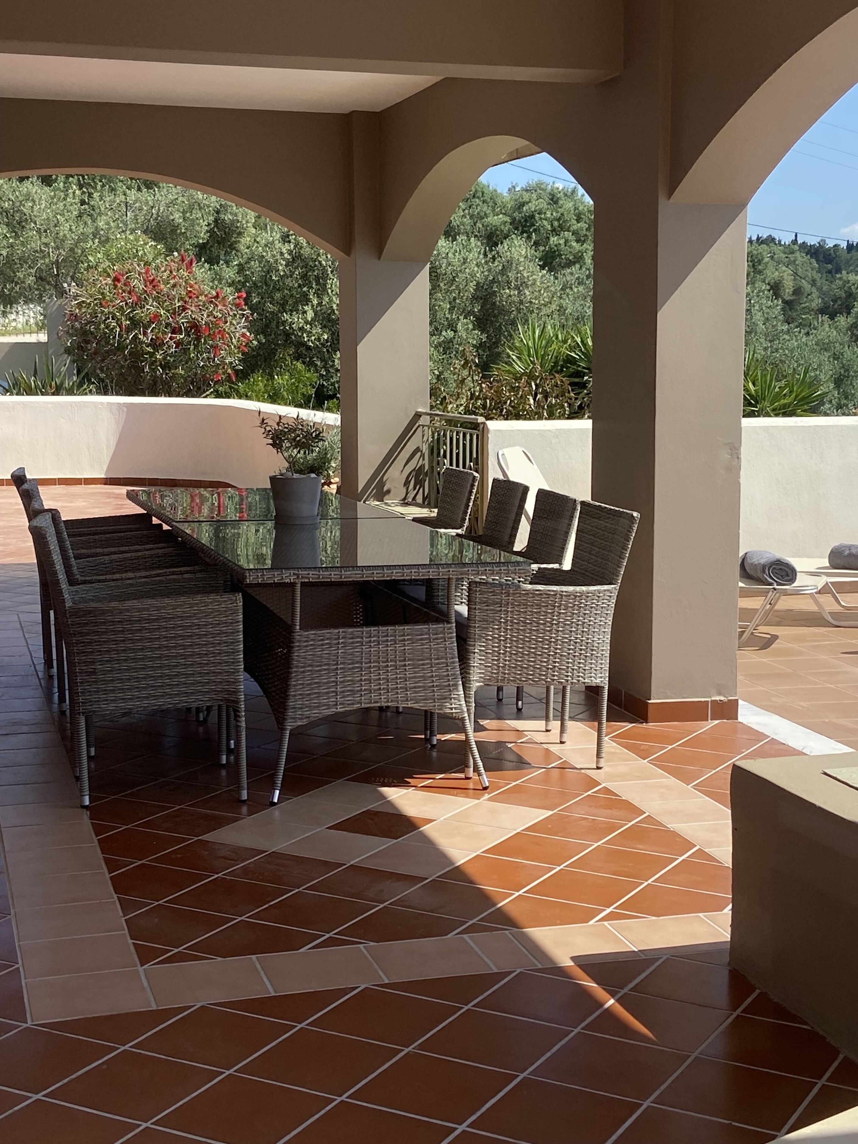 An outdoor patio with a table and chairs, surrounded by a tiled floor and a view of greenery.