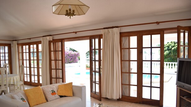 A spacious living room with a white sofa, a glass coffee table, and a television. Large windows with curtains provide a view of a pool and greenery outside. - House / Villa with private pool (San Miguel de Abona)