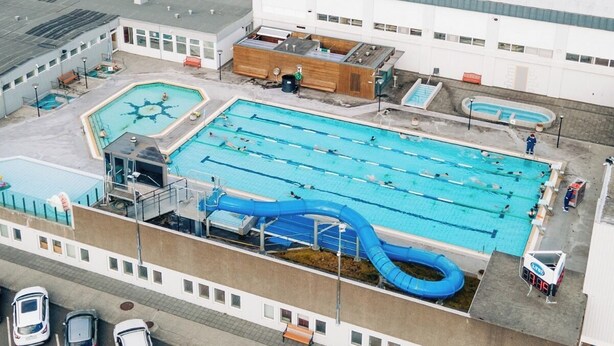 An aerial view of a swimming pool complex with multiple pools, a blue water slide, and a building with a flat roof.