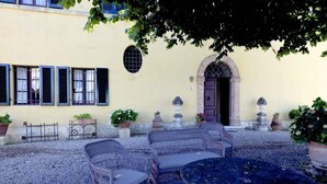 An outdoor seating area with wicker furniture and a round table, surrounded by potted plants and a building with a yellow facade and a brick archway. - Villa with unique view of Siena with private pool (Ascarello)