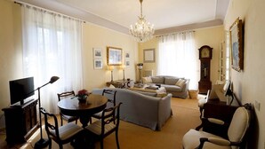 A living room with a sofa, a dining table with chairs, a TV, a chandelier, and a clock on the wall. - Villa with unique view of Siena with private pool (Ascarello)
