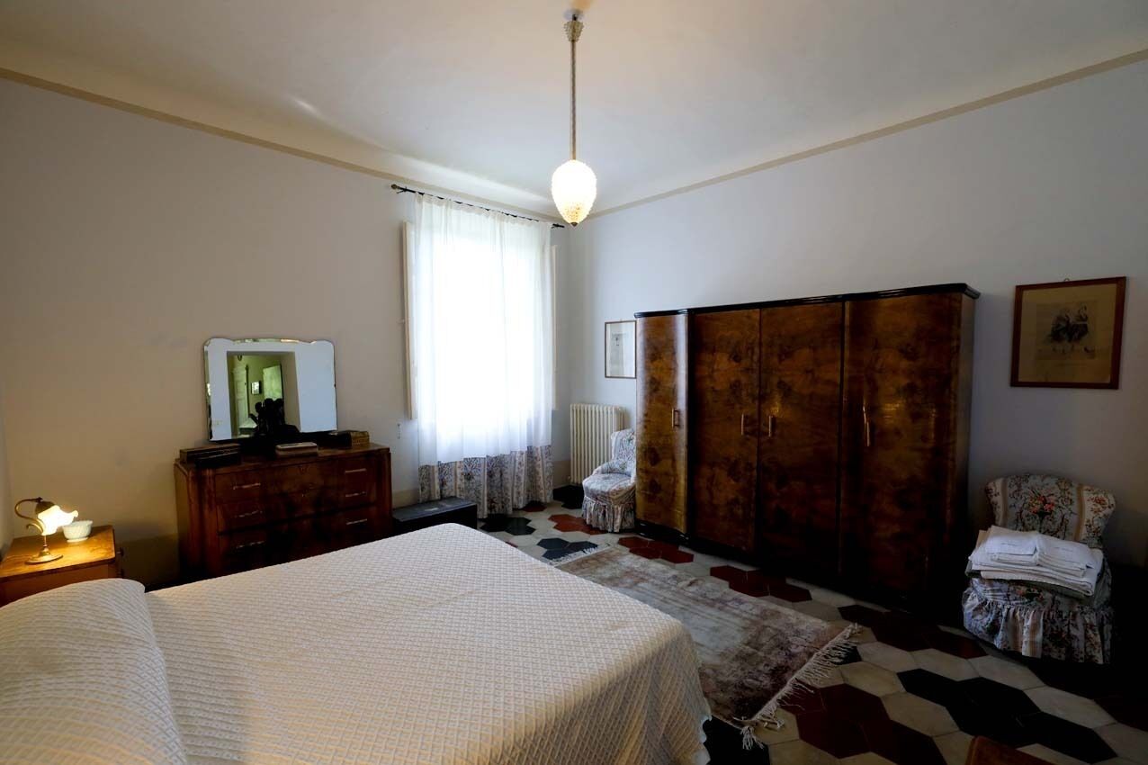 A bedroom with a bed, a wooden wardrobe, a chair, a radiator, and a mirror on a dresser.