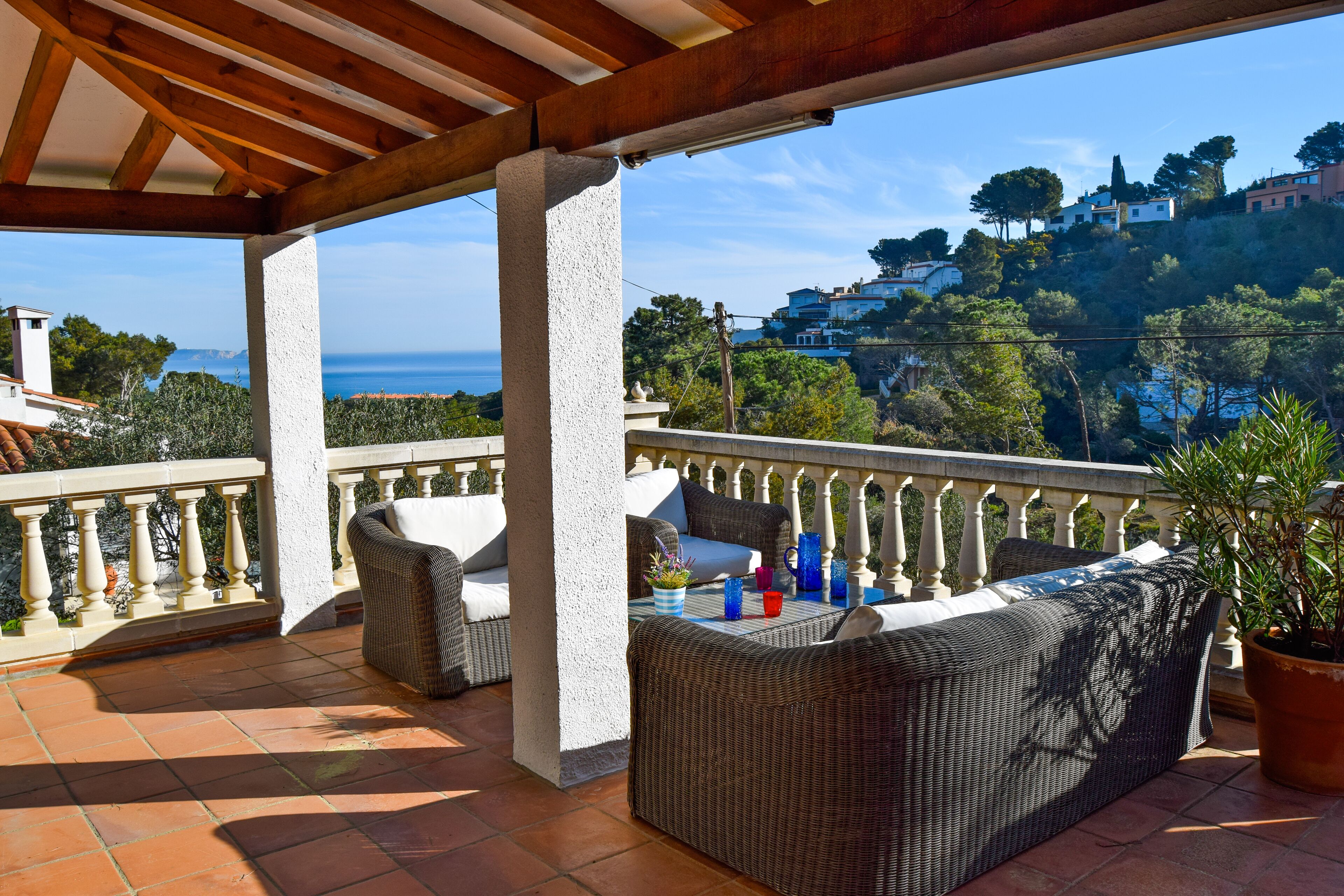 A covered outdoor seating area with wicker furniture and a view of a hillside with buildings.