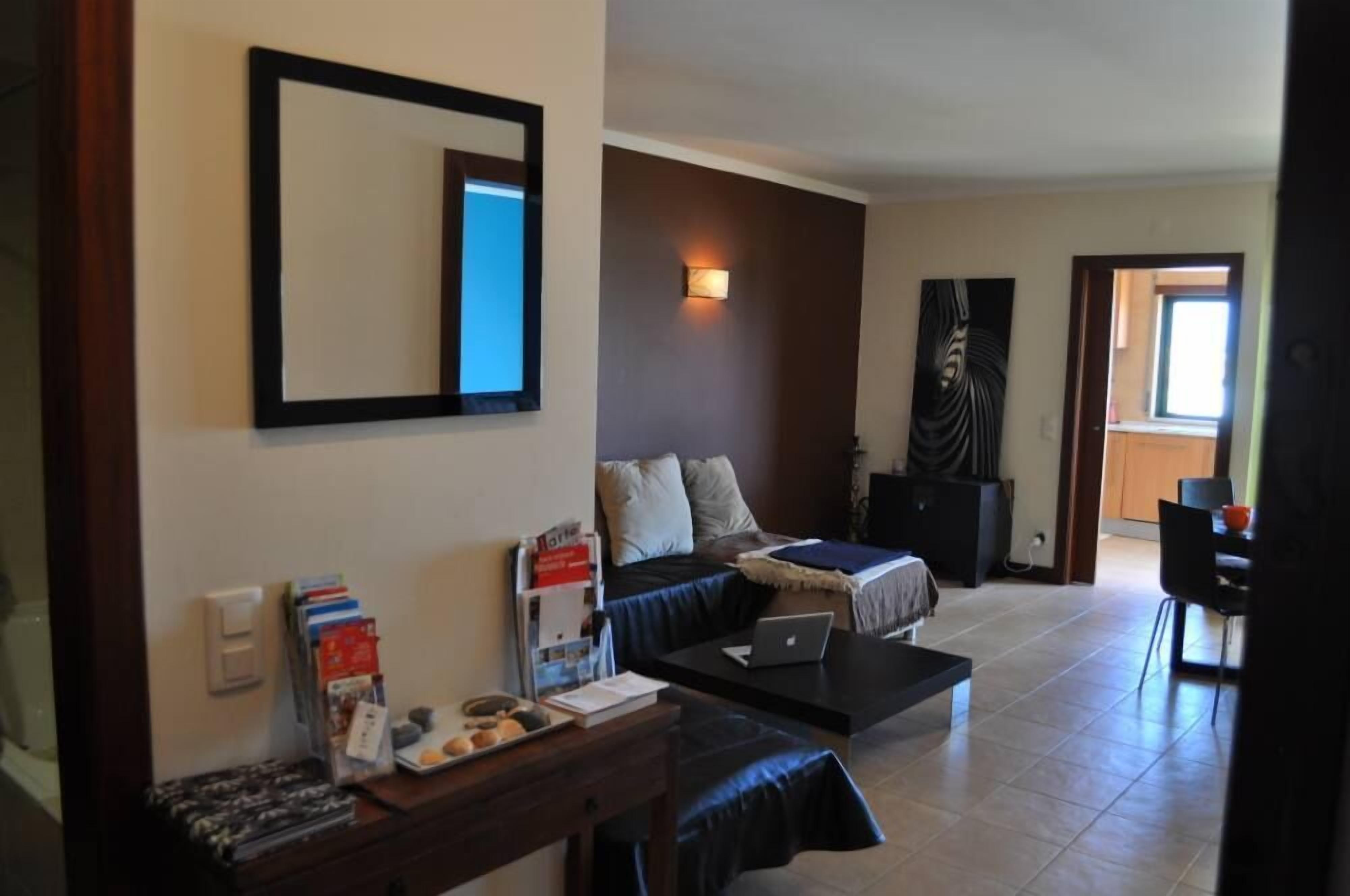 A living room with a sofa, coffee table, and a laptop. There is a bookshelf with books and a small table with a laptop.