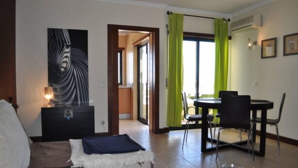 A living room with a sofa, a coffee table, and a dining area with a table and chairs. There is a painting on the wall and a window with curtains. -  Apartment sea view in Salgados Village (Guia)