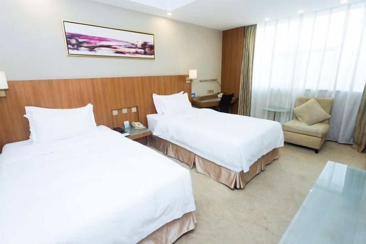 Quarto de hotel com duas camas, uma escrivaninha, uma cadeira e uma janela grande com cortinas.