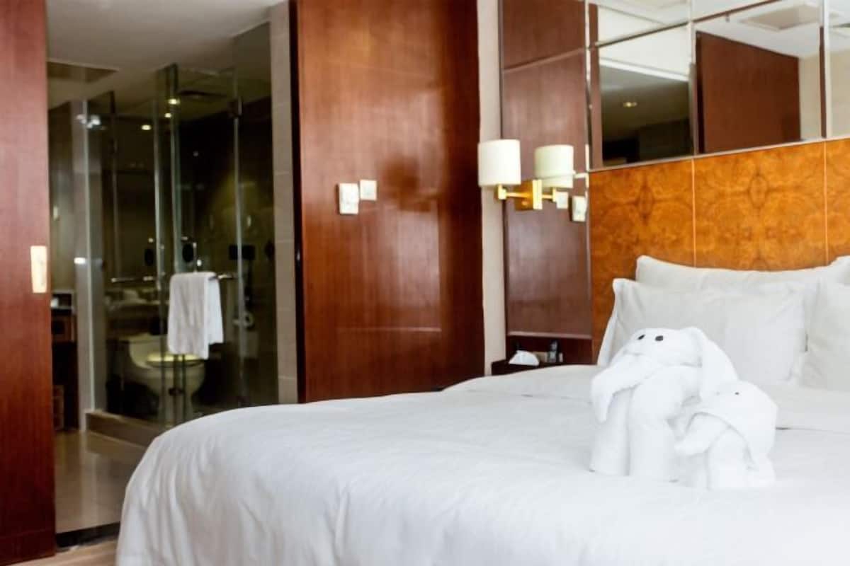 Quarto de hotel com uma cama grande, cabeceira de madeira, box de chuveiro em vidro e um espelho.