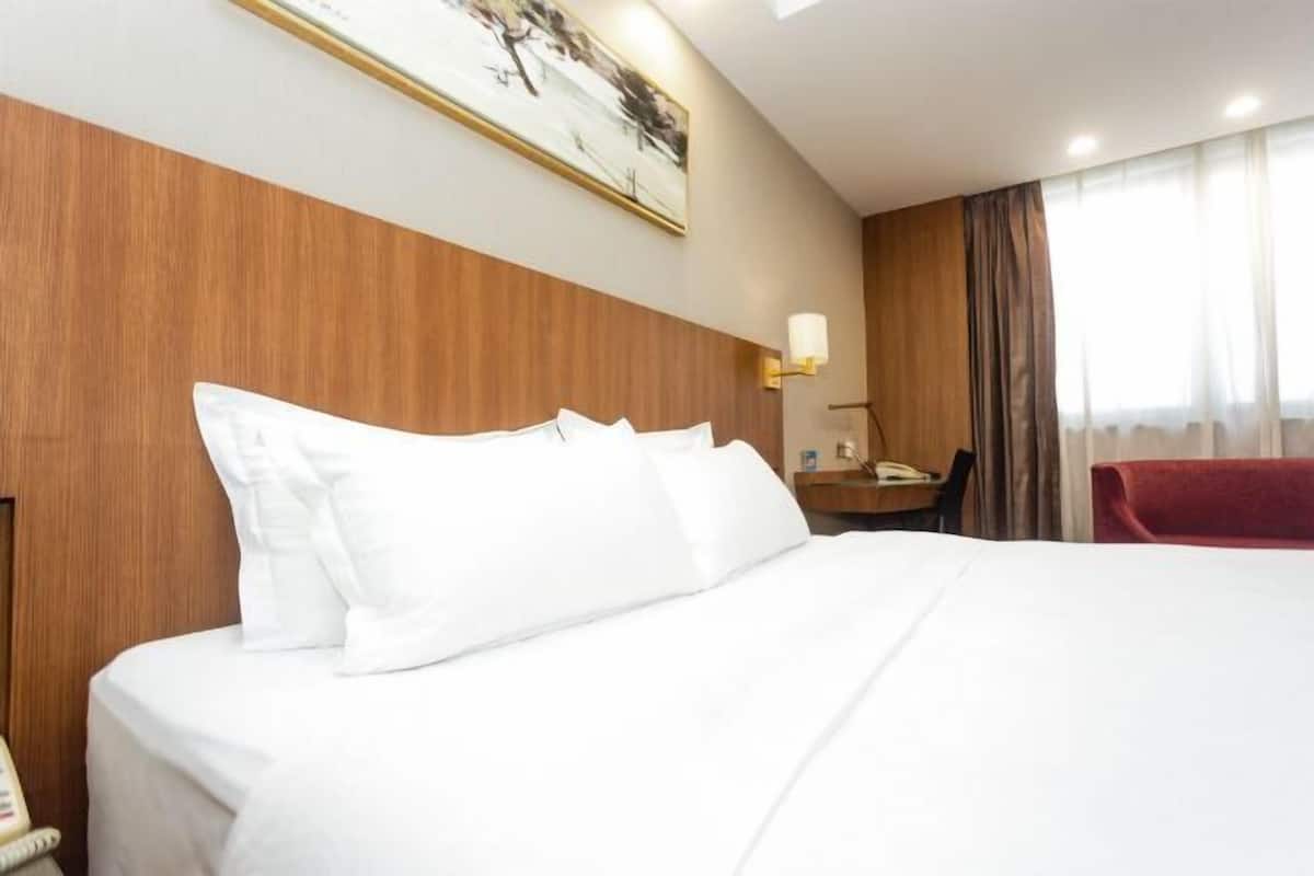 Quarto de hotel com uma cama grande, cabeceira de madeira, escrivaninha com luminária e uma cadeira vermelha.