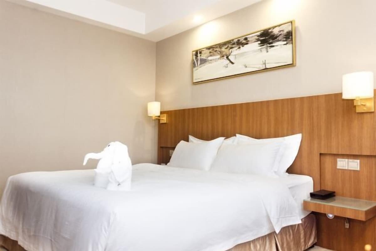 A hotel room with a large bed, wooden headboard, two bedside lamps, a framed picture on the wall, and a small table with a book.