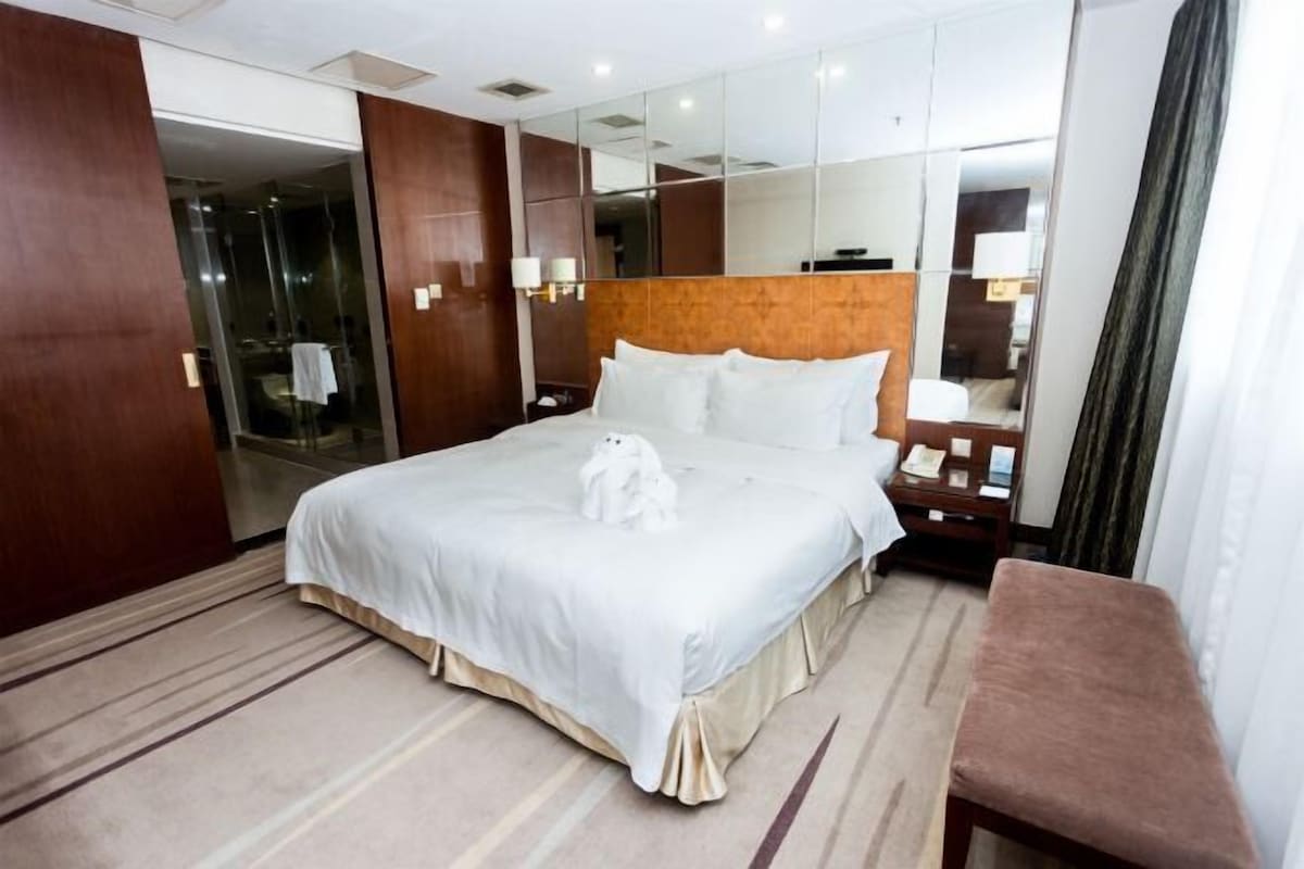 A hotel room with a large bed, a bedside table, a mirror, and a bench.