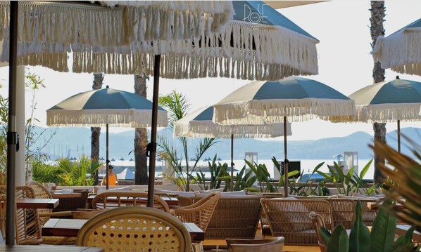 Outdoor seating with umbrellas and wicker furniture, overlooking a body of water and mountains.