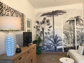 A room with a large mural of tropical foliage, a wooden cabinet with a lamp, a television, and a round table.