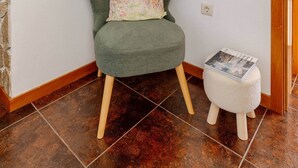 A room with a green armchair, a small stool, a mirror, and a tiled floor.