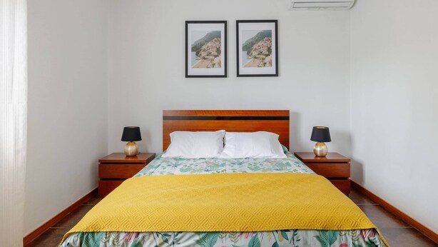 A bedroom with a wooden headboard, a bed with a yellow quilt, two bedside tables with lamps, and two framed pictures on the wall.