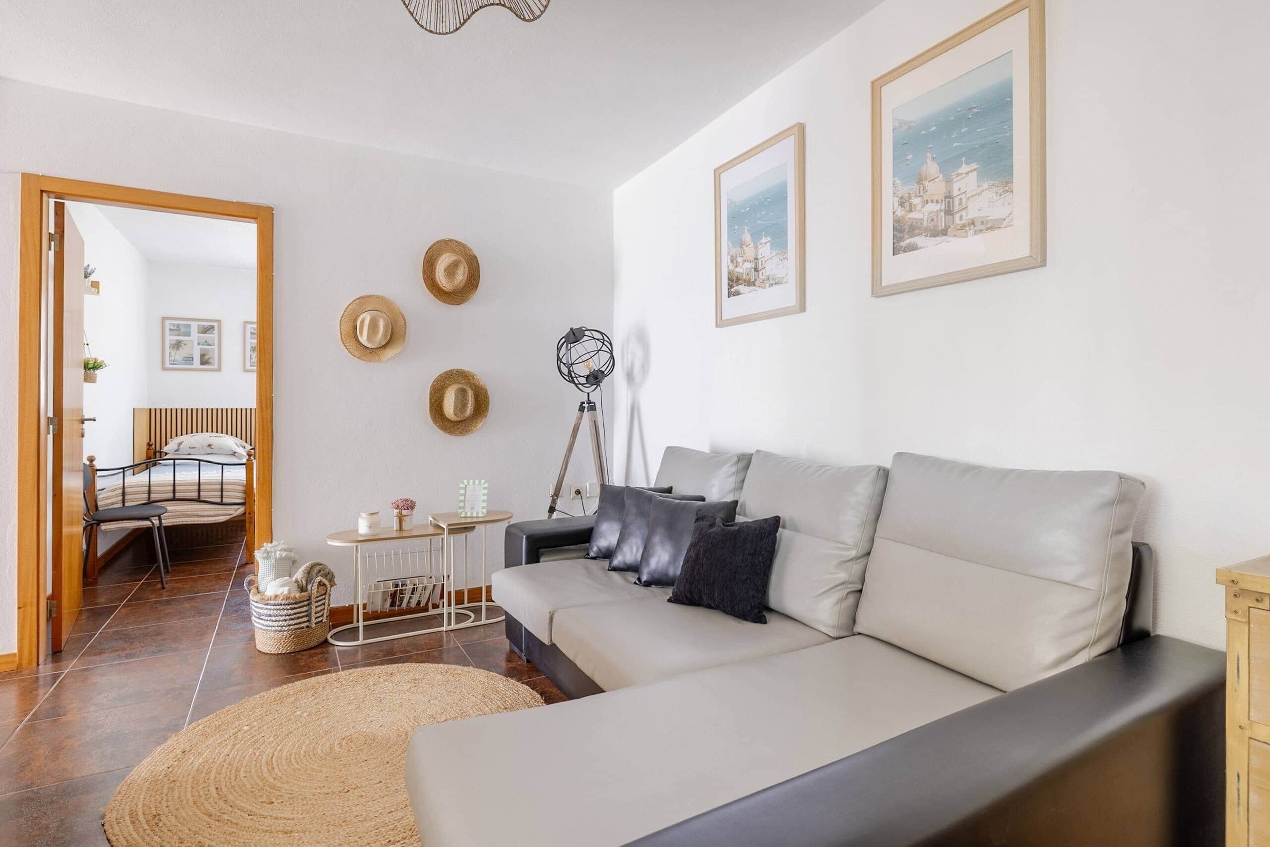 A modern living room with a sofa, a dining table, and a lamp. There are framed pictures on the wall and a door leading to another room.