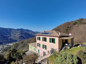 A two-story building with a sloped roof, solar panels, and a satellite dish. It is surrounded by lush greenery and mountains in the background.