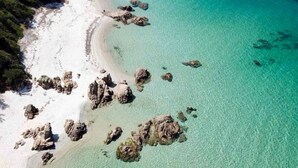 Une vue aérienne d’une plage aux eaux turquoise et limpides, au sable blanc et parsemée de rochers.