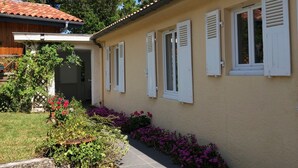 Un edificio su due piani con tetto in tegole e persiane bianche. Davanti all'edificio si estende un giardino con diverse piante e fiori.